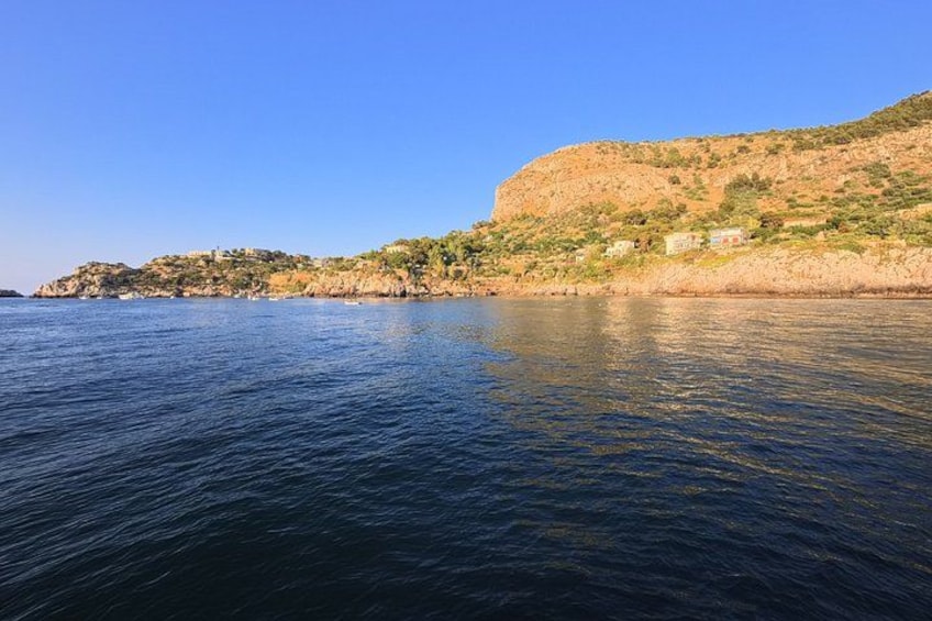 A coastal landscape with calm water, a rocky hill, and a few buildings on the hillside. 3