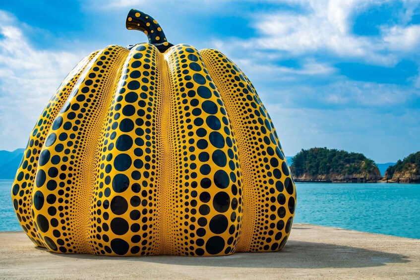 Una escultura grande de color amarillo y negro con lunares, sobre una playa con cielo despejado y agua al fondo. 1
