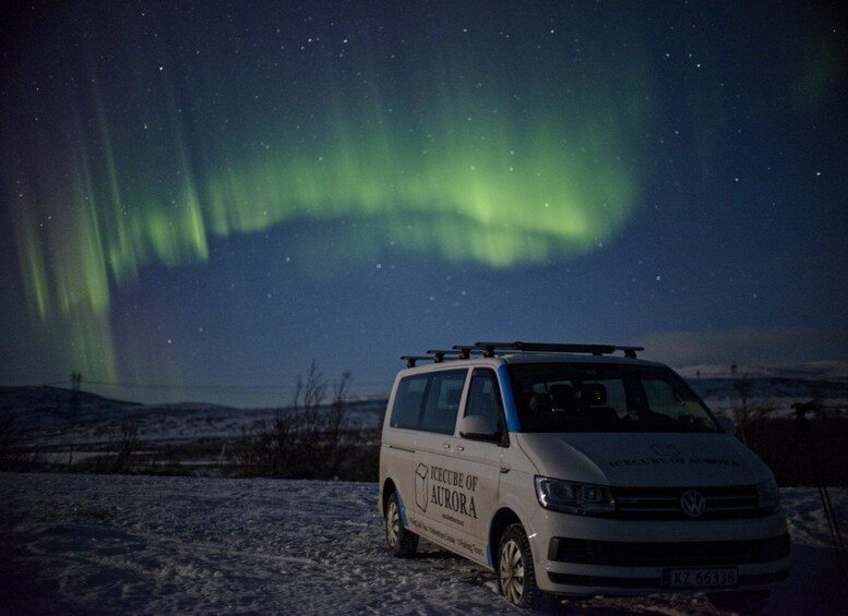 A white van with 'Icecube of Aurora' on its side parked on snow-covered ground under a night sky with the Northern Lights. 2