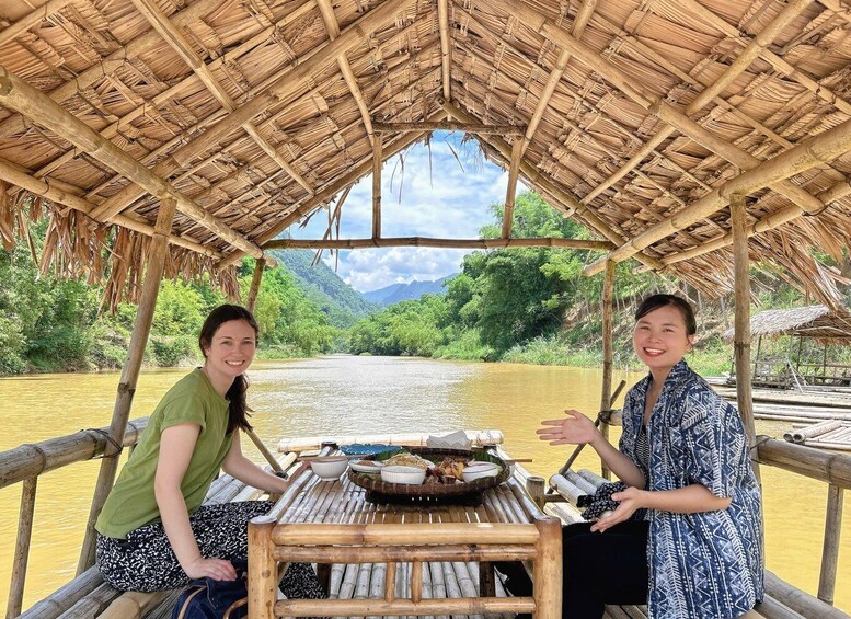 Two people on a bamboo boat with a thatched roof, surrounded by a river and greenery. 4