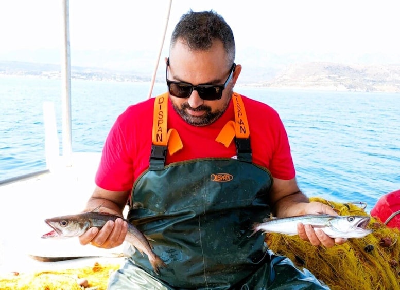 A person holding two fish, wearing a red shirt and a green apron, with a boat and water in the background. 1
