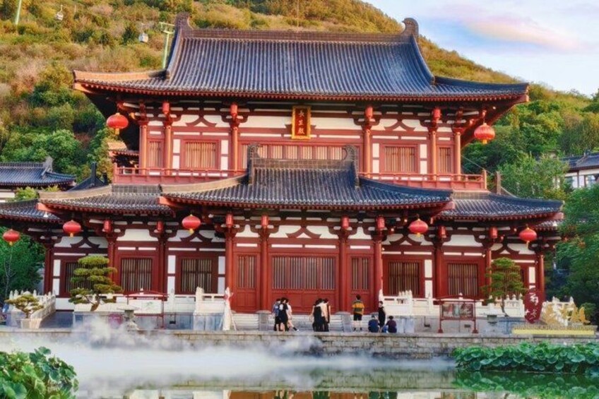 A traditional multi-tiered building with red pillars and a tiled roof, surrounded by greenery and a body of water. 1