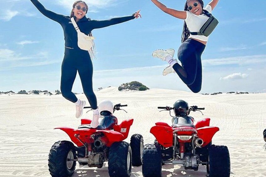 Two people jumping in the air above red ATVs on a sandy beach with a clear sky. 2