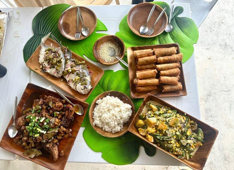 A variety of dishes including fried fish, spring rolls, and a vegetable stir-fry, served on a table with wooden bowls and spoons. 3