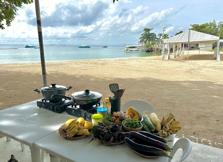 A table with cooking equipment and fresh produce on a beach with boats in the background. 1
