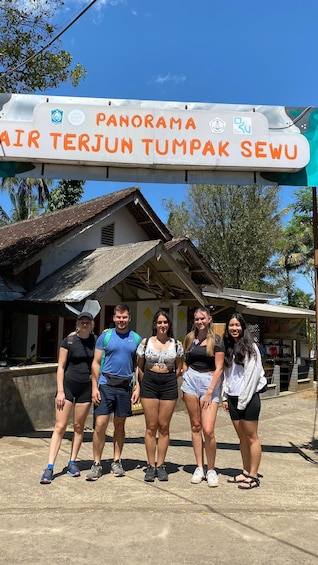 A group of people standing in front of a sign that reads 'PANORAMA AIR TERJUN TUMPAK SEWU' with a rustic building in the background. 5