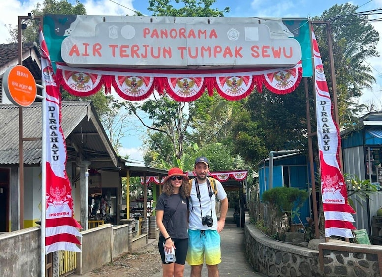 A couple standing under a sign that reads 'PANORAMA AIR TERJUN TUMPAK SEWU' with Indonesian flags and a red and white banner. 2