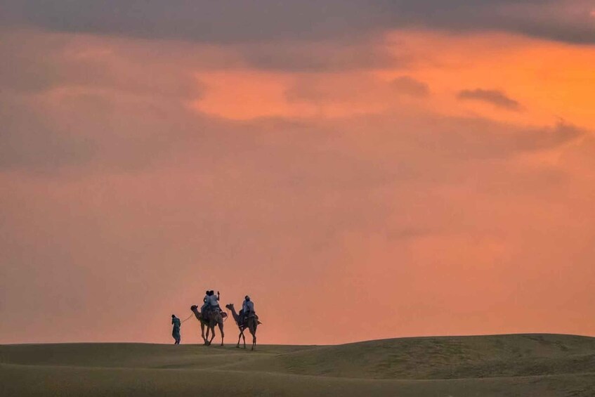 Three people riding camels across a desert landscape during sunset. 4