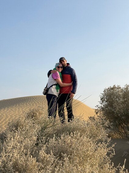 Two people embracing in a desert landscape with sand dunes and sparse vegetation. 3