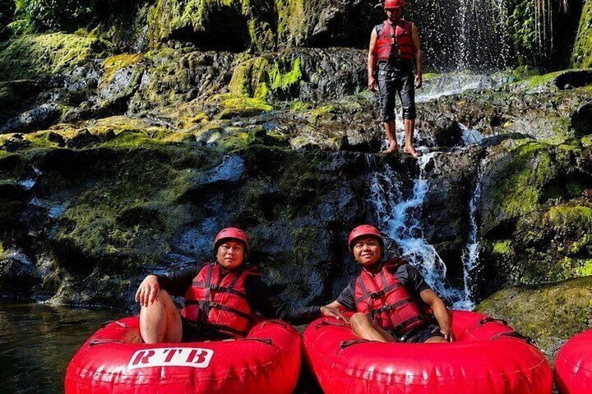 Two people in red life jackets and helmets sitting on red inflatable tubes in a river with a waterfall in the background. 3