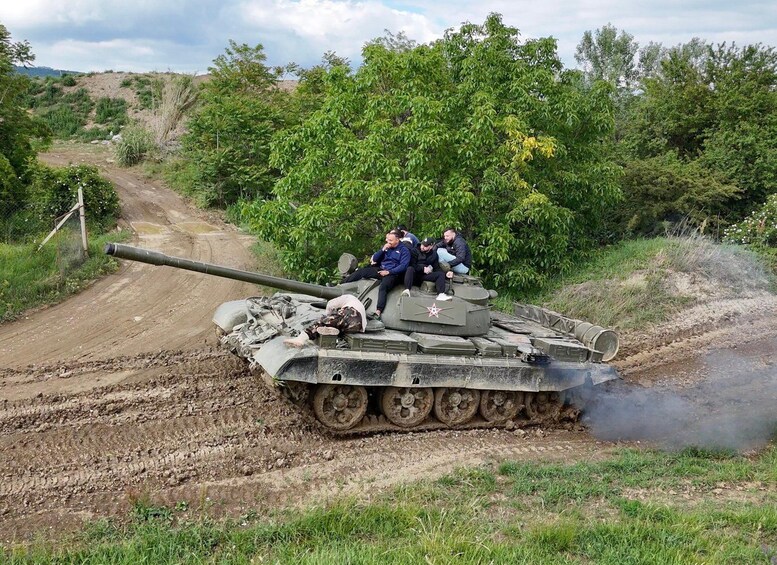 A military tank with soldiers on top, moving on a dirt road surrounded by greenery. 3