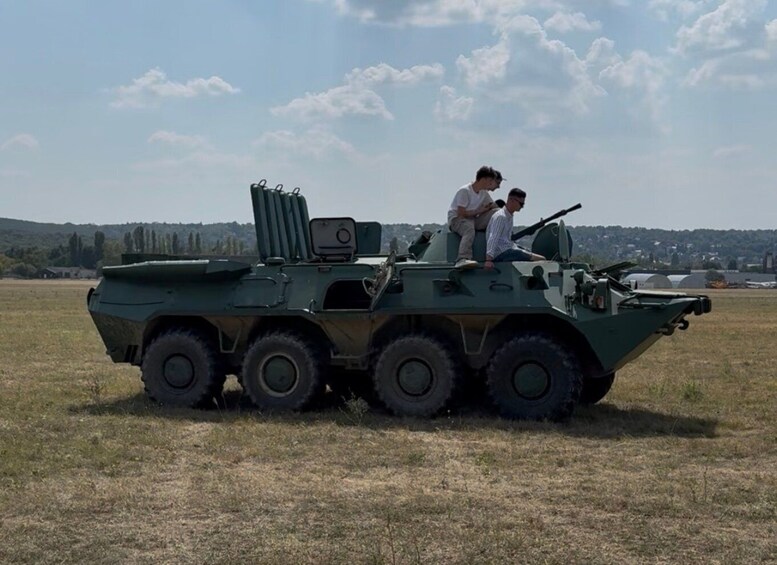 A military vehicle with two people on top, parked in an open field with a distant cityscape. 2