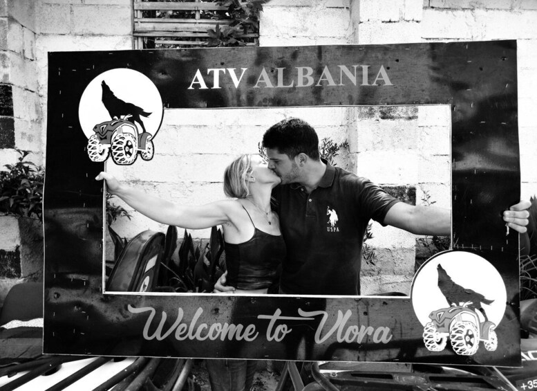 A couple embracing in front of a sign that reads 'ATV ALBANIA Welcome to Vora.' 3