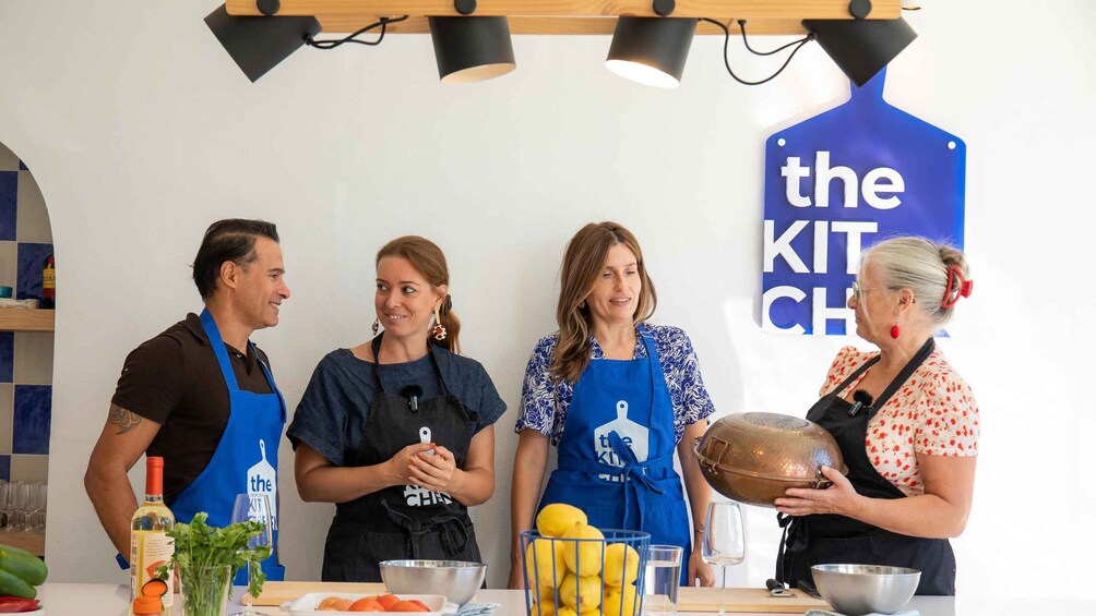 Neljä henkilöä, joilla on esiliinat päällään, seisoo keittiön työtason edessä, ja työtason yläpuolella on kyltti, jossa lukee: ”THE KIT CHEF”. 2