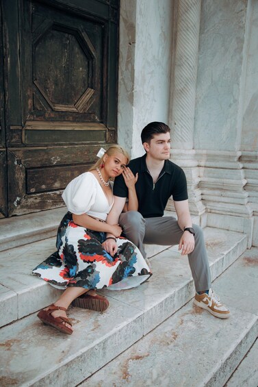 Two people sitting on stone steps with a large wooden door and marble columns in the background. 3