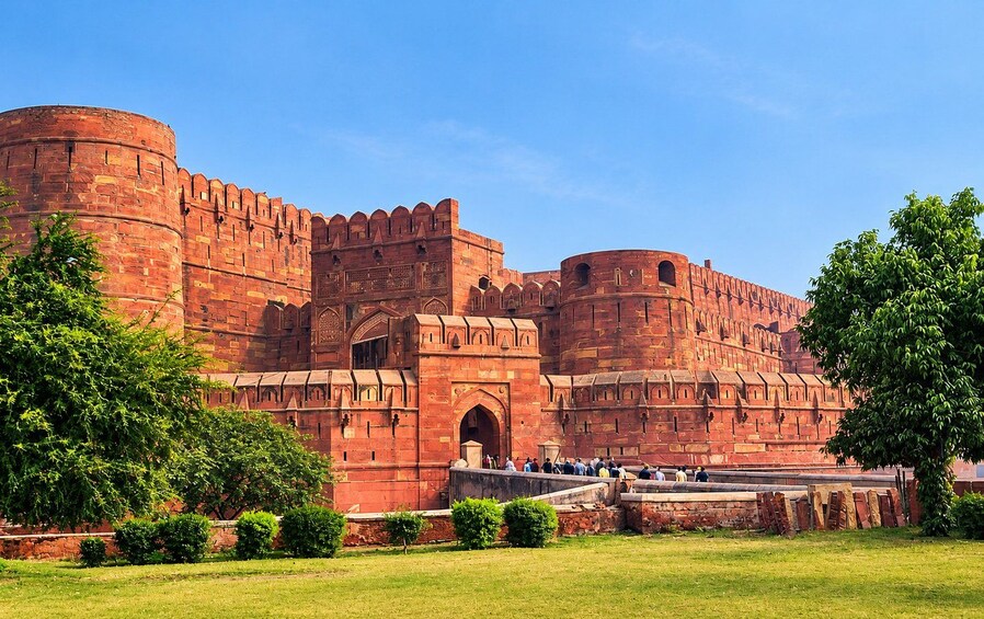 A large red brick fort with multiple towers and arched gateways, surrounded by green trees and grass. 5