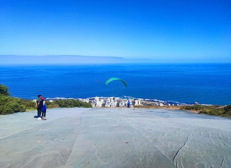 Paesaggio costiero con cielo sereno, una persona in piedi su una superficie in cemento e un paracadute verde in lontananza. 3