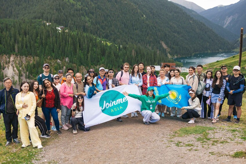 A group of people holding a banner with 'Joinme ASIA' and a flag, standing on a mountain path with a river and forest in the background. 2