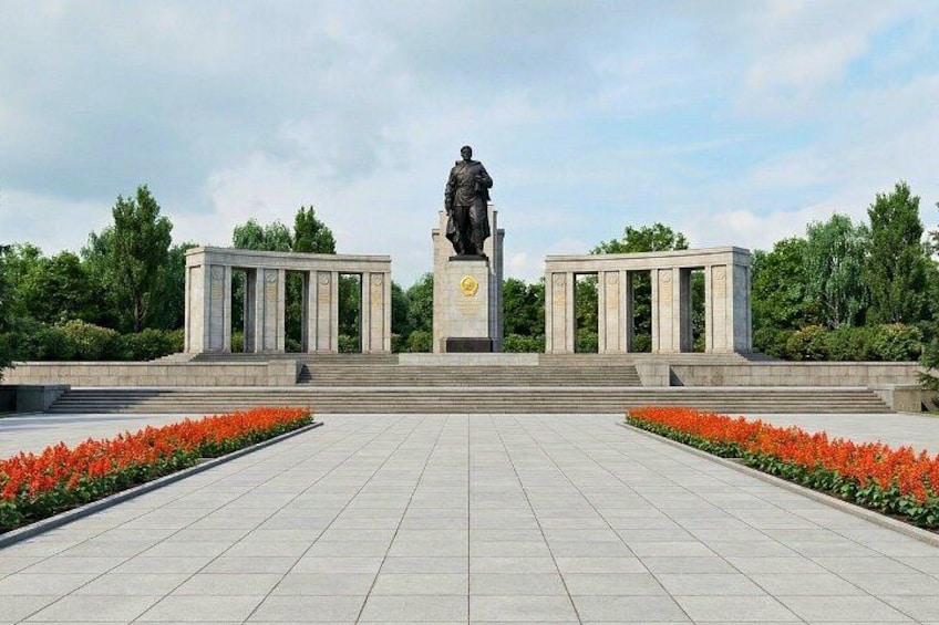 A statue with a pedestal, flanked by two columns, in a landscaped area with red flowers and trees. 2