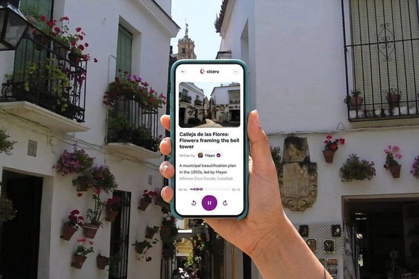 A hand holding a smartphone displaying a photo of a street with flowers and a bell tower. 1