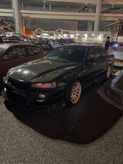 A black car parked in a lot with other vehicles, a pedestrian, and a building in the background. 5