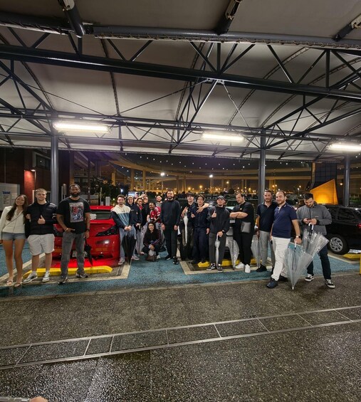 A group of people posing in front of a red sports car in a covered parking area with a metal roof structure. 4