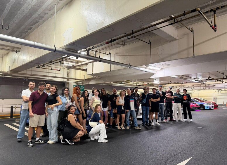 A group of people posing in a parking garage with cars and industrial pipes in the background. 3
