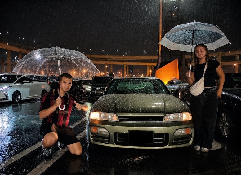 A person crouching in front of a car with a transparent umbrella, another person standing beside a car with a striped umbrella, and a building with lights in the background. 5