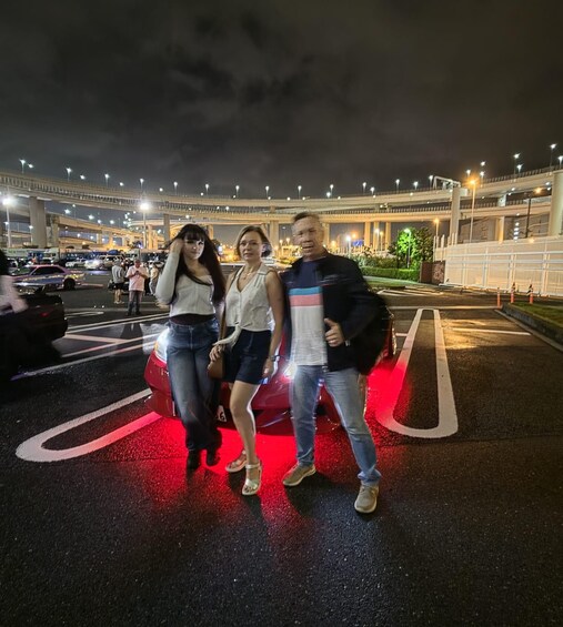 Three people standing on a road with a red line, a bridge, and streetlights in the background. 2