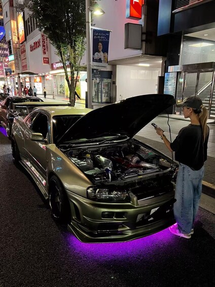 A car with its hood open, parked on a street with purple underglow lights, surrounded by urban buildings and pedestrians. 3