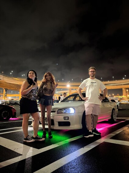 Three people standing in front of a white car with colorful lights on the ground, under a bridge at night. 5