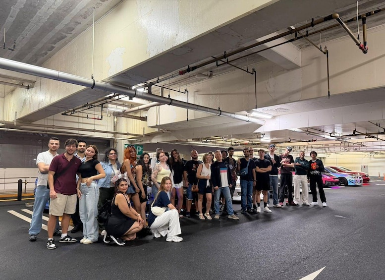 A group of people posing in a parking garage with cars and industrial pipes in the background. 3