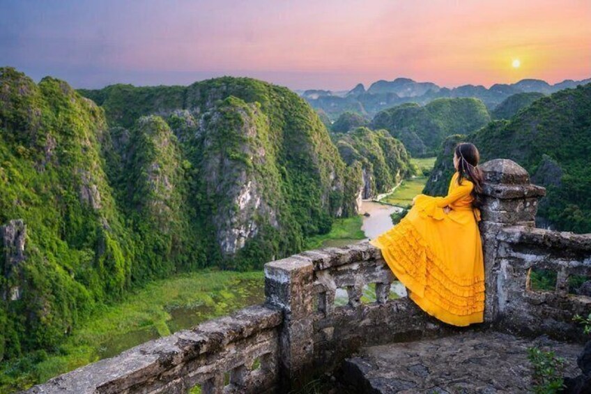 A person in a yellow dress standing on a stone wall overlooking a lush valley with a river. 1