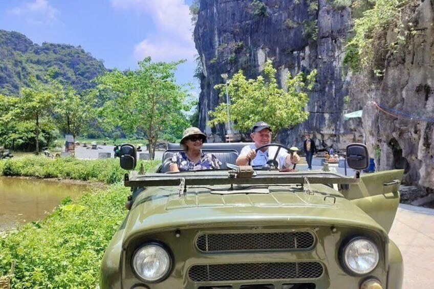 A vintage military vehicle with two passengers, a rocky cliff, and a river in the background. 3