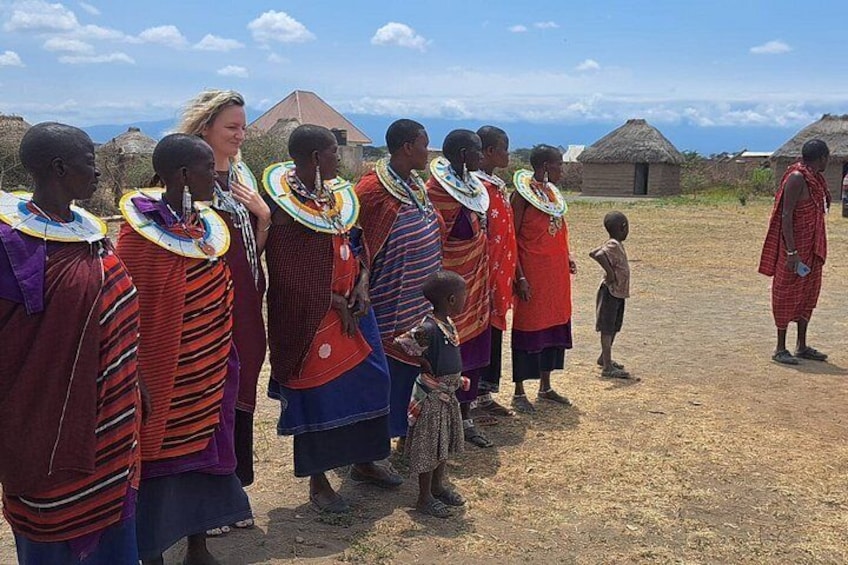 A group of people in traditional attire with beaded necklaces, standing in a line outdoors with thatched-roof huts in the background. 2