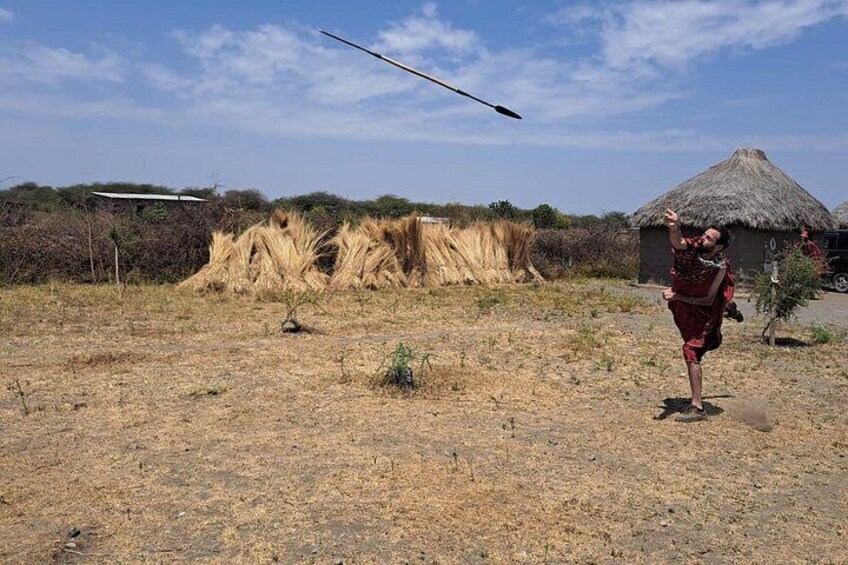 A person in traditional attire throwing a spear in a dry, grassy field with thatched-roof huts in the background. 4