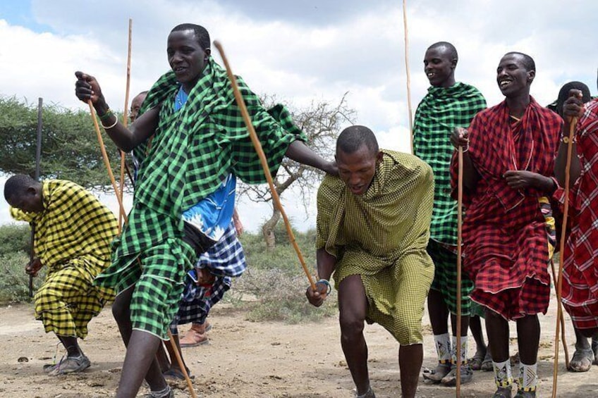 A group of people in traditional attire, some holding sticks, in an outdoor setting with sparse vegetation. 1