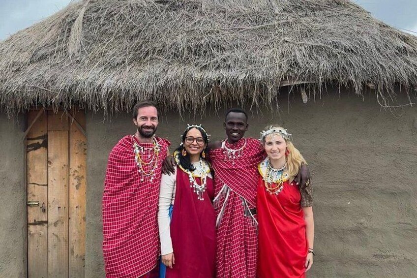 Four people in traditional attire standing in front of a thatched-roof hut. 5
