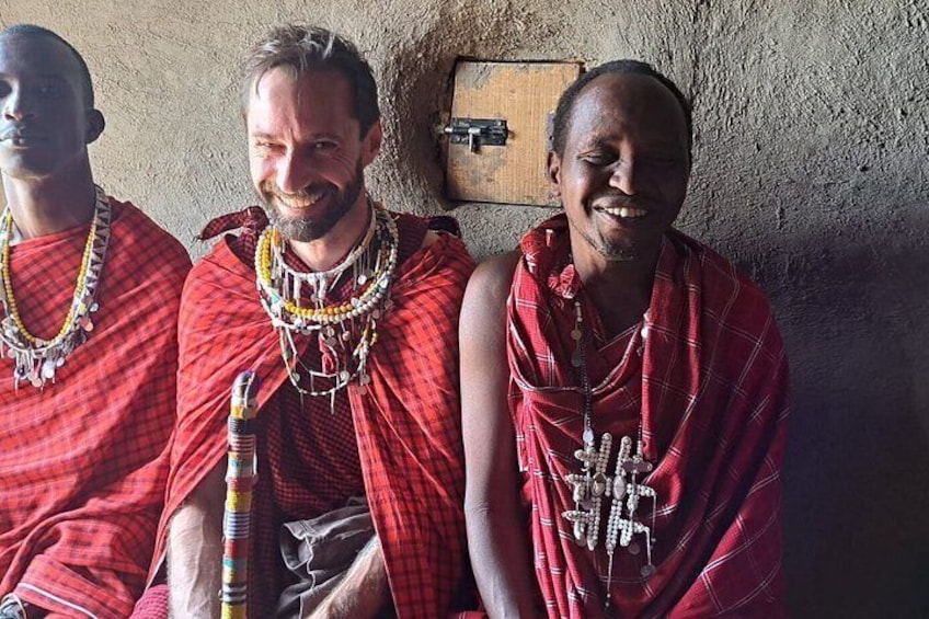 Three individuals wearing traditional red garments and beaded necklaces, seated against a textured wall. 3
