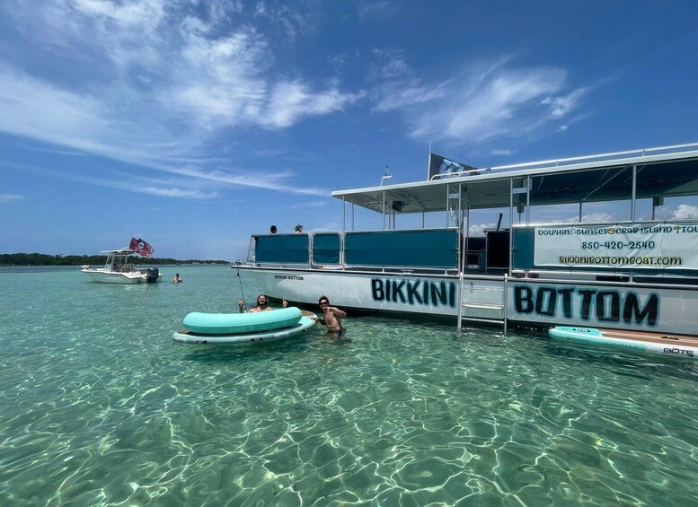 A clear blue sea with a boat named 'Bikkini Bottom' docked, people on inflatable rafts, and another boat with a flag in the background. 4