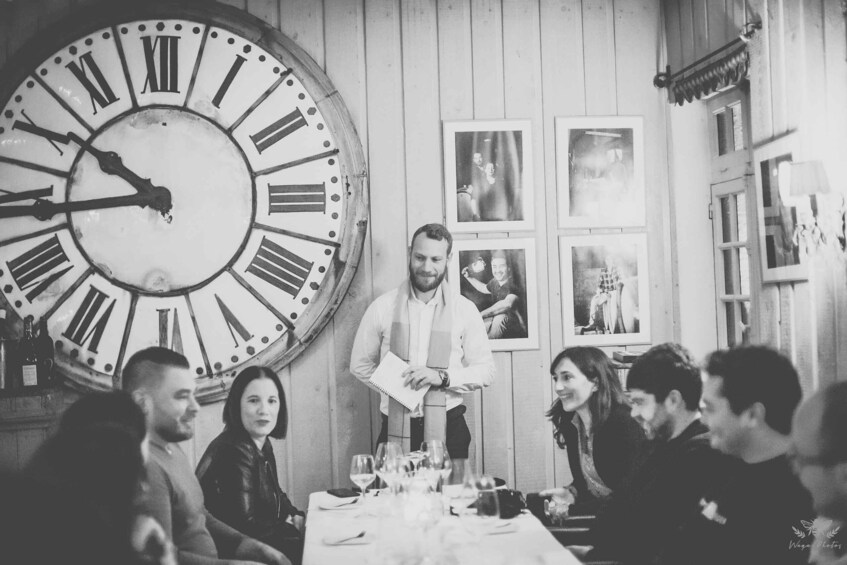 A group of people seated around a table with a large clock and framed pictures on the wall. 1