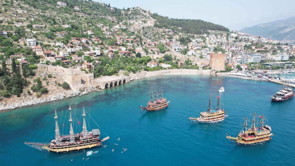 A coastal town with historic architecture, a fortified wall, and several traditional boats anchored in the bay. 3