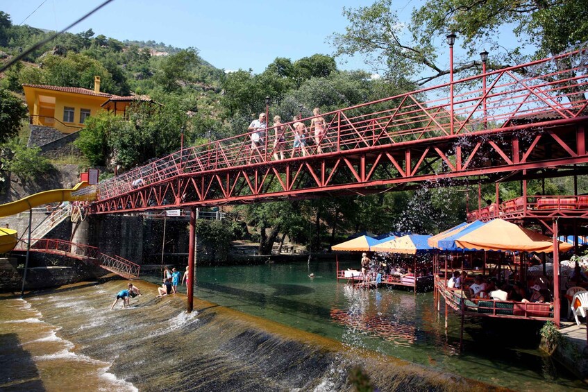 A red metal bridge over a river with people walking, a water slide, and a boat with passengers. 4