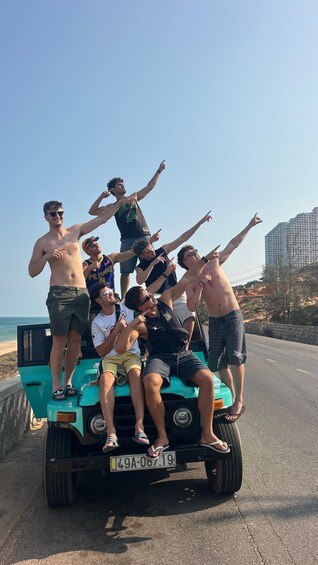 A group of people on a teal vehicle with a license plate '49A-087-19' by a coastal road with buildings in the background. 3