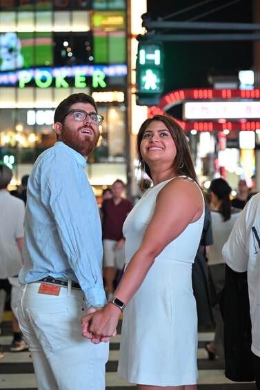 A couple holding hands in a busy urban area with neon signs and a pedestrian crossing. 4