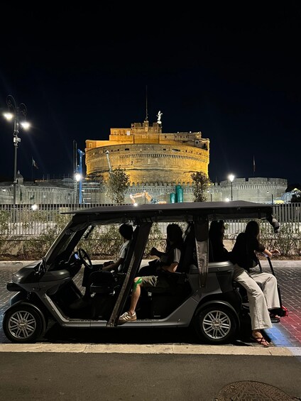A golf cart with three passengers parked on a street at night, with a historic building illuminated in the background. 2