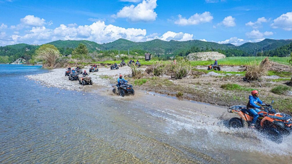 People riding ATVs across a shallow river, surrounded by green hills and a clear sky. 4