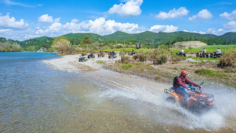 A riverbank with people riding ATVs, a clear sky, and green hills in the background. 5