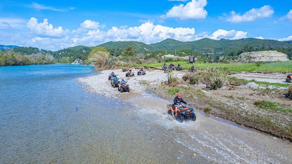 People riding ATVs along a riverbank with mountains in the background. 3