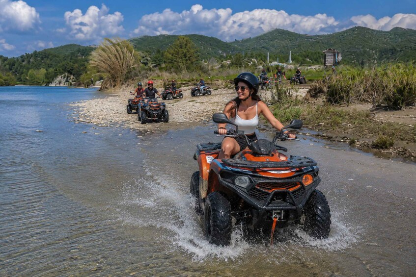 A person riding an ATV through shallow water, with a group of ATVs and riders in the background on a riverbank with mountains and greenery. 1